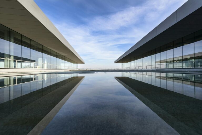 Futuristic glass building mirrored on a still water surface with a cloudy sky.